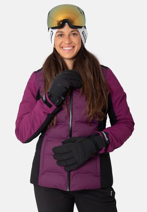 Lila und schwarze, gefütterte Skijacke mit einem Reißverschluss, kombiniert mit schwarzen Handschuhen. Das Modell trägt Skibrillen und ein weißes gestricktes Stirnband.