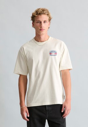 Jeune homme aux cheveux blonds portant un T-shirt crème avec un petit logo circulaire rouge et bleu sur la poitrine gauche et un pantalon noir.