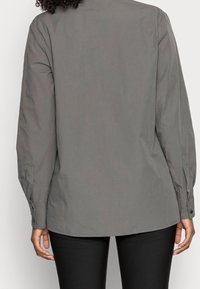 Camicia grigia con bottoni e maniche lunghe, con yoke posteriore e polsini con dettagli a bottone. Il tessuto appare liscio con texture minima.