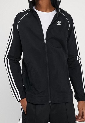 Mężczyzna noszący czarną kurtkę dresową Adidas z białymi paskami na rękawach i białym logo Adidas na piersi, w połączeniu z czarnymi spodniami.
