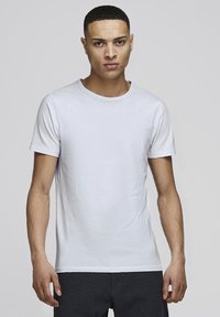 Man with short hair wearing plain light gray short-sleeve t-shirt and dark textured pants, standing against a plain light background.