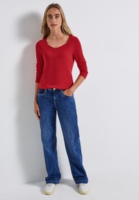 Rotes Langarmoberteil mit V-Ausschnitt, kombiniert mit blauen geraden Jeans. Weiße Sneaker runden das Outfit vor einem schlichten Hintergrund ab.