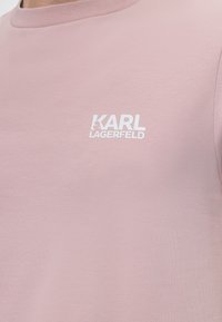 Κοντινό πλάνο ενός ανοιχτό ροζ T-shirt με λευκό λογότυπο "KARL LAGERFELD" τυπωμένο στο στήθος.