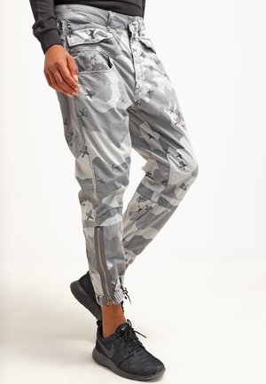 Personne portant un pantalon cargo camouflage gris et blanc avec des fermetures éclair latérales et des chaussures de sport noires, debout avec une jambe croisée.