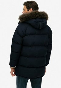 Chaqueta de plumas azul marino con capucha forrada de piel, que presenta una textura acolchada y un corte más largo, vista desde atrás con detalles de costuras visibles.