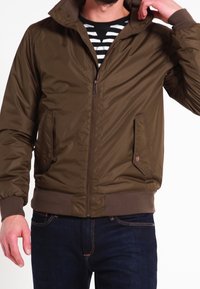 Veste bomber matelassée marron avec un col montant, une fermeture éclair, des poches latérales et des poignets côtelés ; portée avec une chemise rayée en dessous.