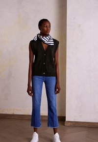 Colete malha verde-oliva com grandes botões, combinado com jeans claros de perna larga e ténis brancos. Acessório com um lenço riscado.