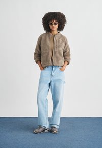 Plišani smeđi shearling jakna s ovratnikom, bež top, široke svijetloplave traperice i sive tenisice; model stoji s rukama u džepovima.