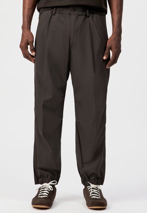 Mężczyzna w luźnych czarnych spodniach z elastycznymi mankietami i ciemnych trampkach z białymi sznurówkami, stojący na jednolitym tle.