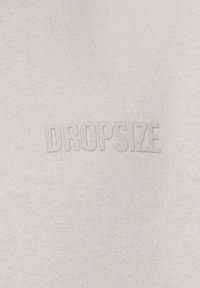 Svijetlosiva tkanina s reljefnim tekstom "DROPSIZE". Materijal izgleda glatko s suptilnom teksturom. Nema dodatnih uzoraka ili akcenta.