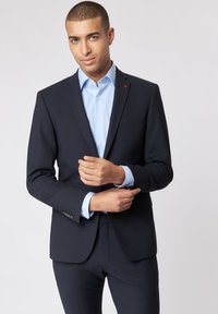 Granatowa marynarka o usztywnionej konstrukcji, z klapą typu notch, jedną kieszenią na piersi oraz czerwonym akcentem na kołnierzu. Pod spodem jasnoniebieska koszula.