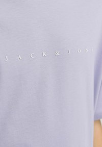 Šviesiai purpurinė marškinėliai, pagaminti iš minkštos medvilnės, su baltu logotipu "JACK & JONES", atspausdintu per krūtinę minimalistiniu šriftu.