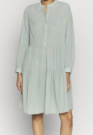 Kobieta w luźnej, długorękawowej, jasnizielonej sukience z falbanami, z guzikami przy piersi i okrągłym kołnierzykiem.