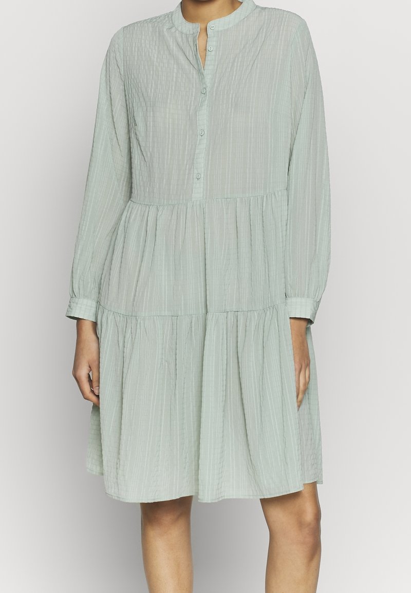 Femme portant une robe ample à manches longues, vert clair, à volants, avec des boutons sur la poitrine et un col rond.