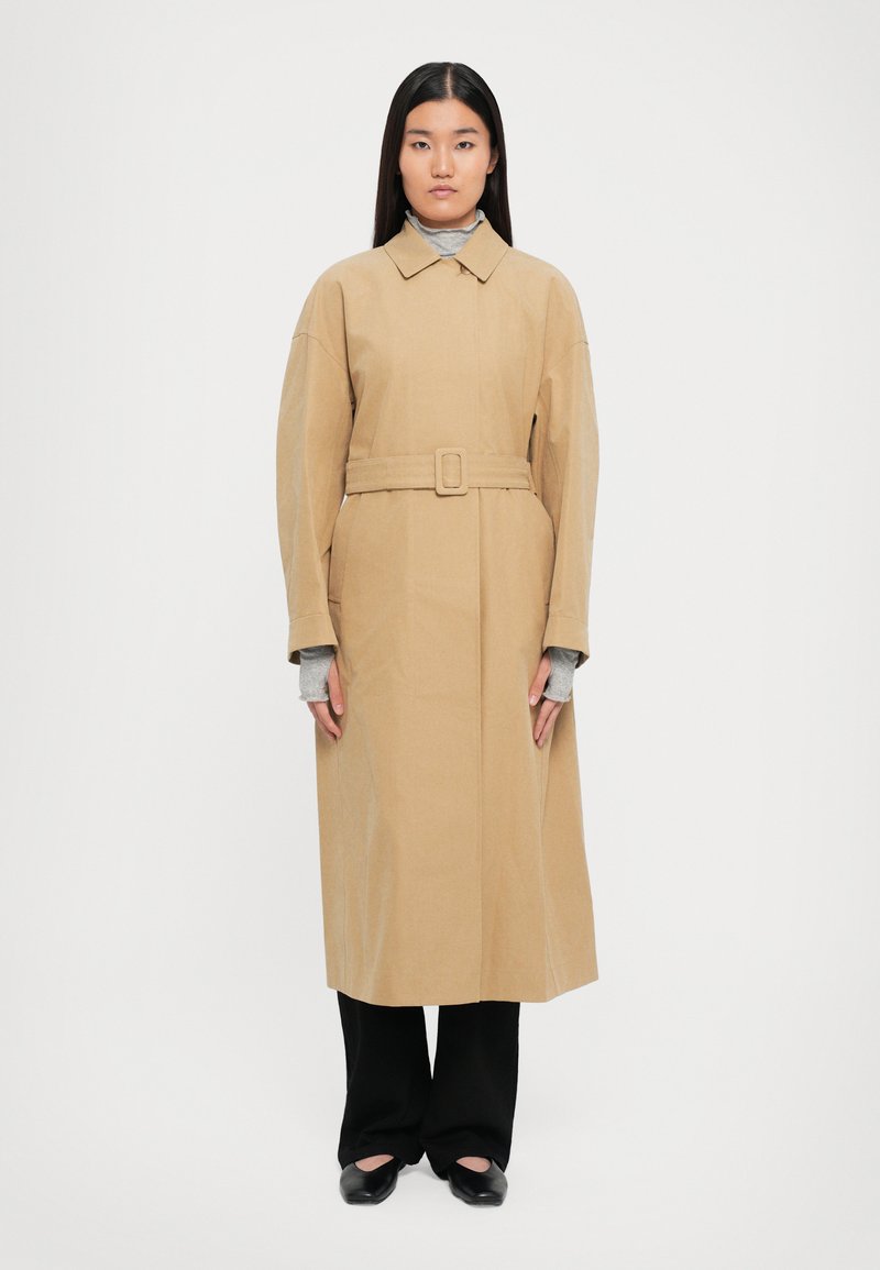 En kvinna står framåt med en beige knyttrenchcoat över grå långärmade tröjor, svarta byxor och svarta skor mot en vit bakgrund.