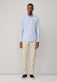 Camicia button-up azzurro chiaro con righe verticali, dotata di taschino verde sul petto. Abbinata a pantaloni beige e scarpe scure.