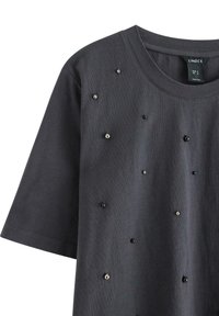 Mørkegrå t-shirt lavet af bomuld, med korte ærmer og pyntet med forskellige størrelser af sorte og sølvfarvede nitter over fronten.