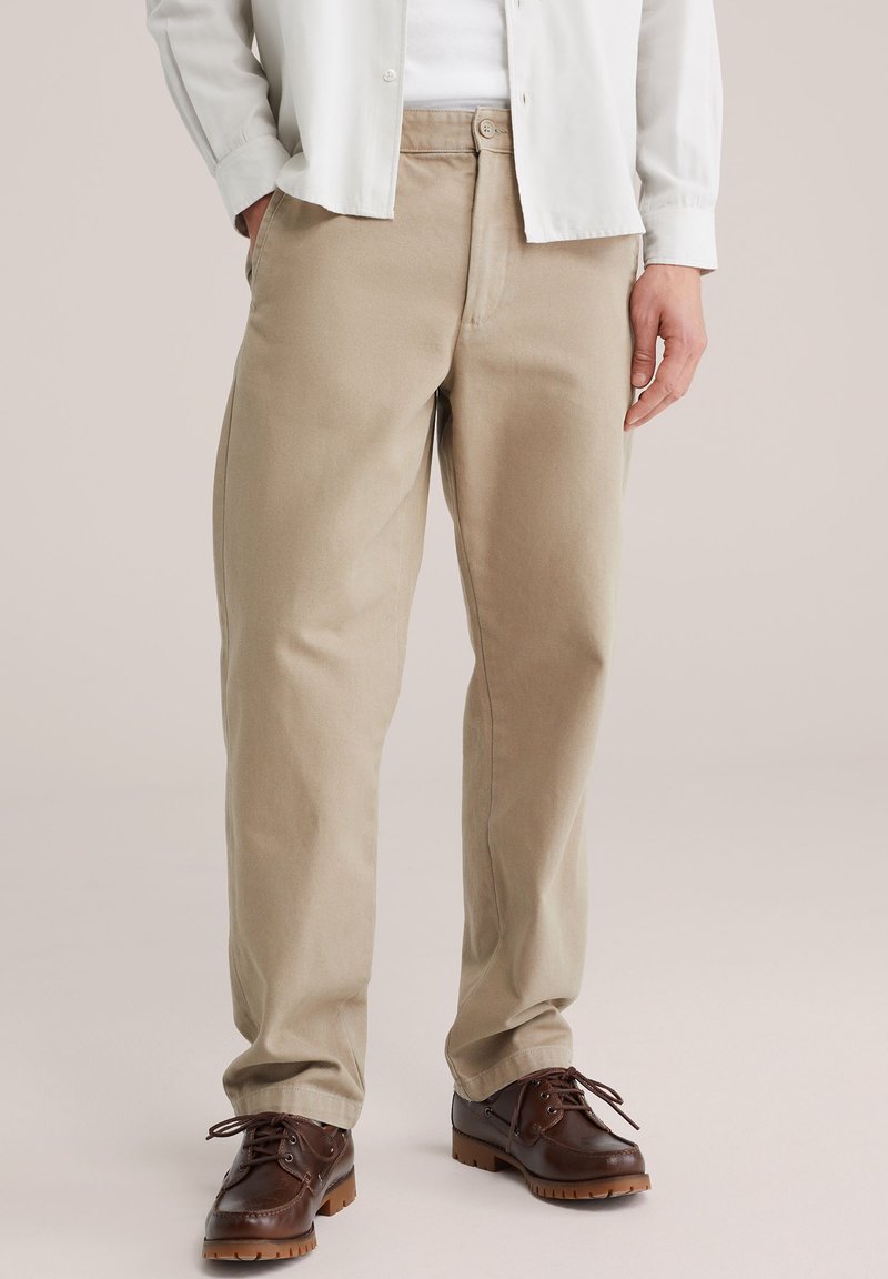 Muž vo svetlohnedých nohaviciach s rovnými strihmi, bielej košeli a tmavohnedých kožených topánkach na šnurovanie stojaci na svetlom pozadí.