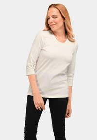 Lichtbeige langesleeve shirt met een ronde halslijn, voorzien van een subtiele bies. Het materiaal lijkt glad en rekbaar, gecombineerd met donkere broeken.