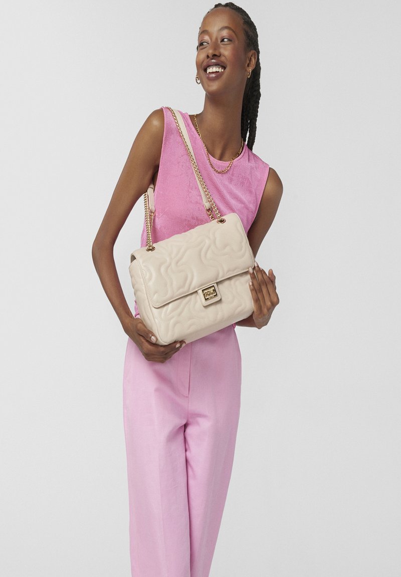 Bolso de mano beige acolchado con solapa plegable, correa de cadena y acentos de hardware dorado. Textura suave y detalle de patrón ondulado.