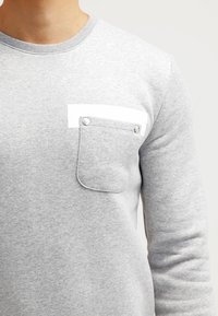 Felpa grigia con scollo rotondo, caratterizzata da una tasca decorativa bianca con bottoni a pressione, tessuto testurizzato e maniche lunghe.