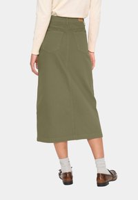Falda midi verde oliva de algodón con corte recto, que presenta dos bolsillos traseros y una cinturilla. Se lleva con calcetines gris claro y zapatos marrones.