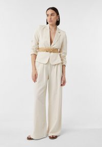 Jasno beżowa marynarka dopasowana z głębokim dekoltem w kształcie litery V, w zestawieniu z spodniami o szerokich nogawkach i plecionym paskiem ze słomy. Prosty krój, gładka tekstura, minimalne akcenty.