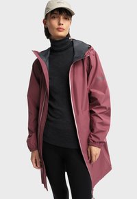 Mlada osoba u prašnjavopinkom kišnom kaputu s kapuljačom, ispod kojeg nosi tamni dolčevita i crne hlače, te bež kapu.