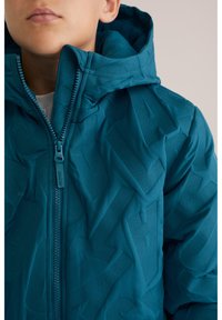 Chaqueta acolchada color teal con collar alto, que presenta un patrón de texto en relieve. El cierre frontal con cremallera y la tela texturizada acentúan el diseño.