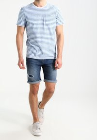 Man som går, iklädd en ljusblå och vit randig kortärmad skjorta, uppvikta mörka jeansshorts med revor, och vita sneakers.