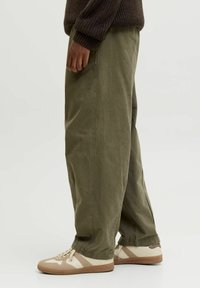 Pantaloni a gamba larga color verde oliva realizzati in tessuto morbido, abbinati a sneakers beige e crema con dettagli testurizzati e design a profilo basso.