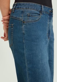 Vaqueros de mezclilla azul con un ligero desgaste, diseño de cinco bolsillos y costuras en contraste. La cintura incluye cierre de botón y cremallera.