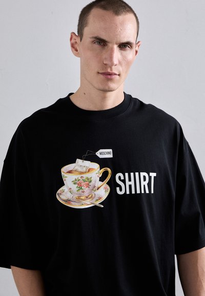 T-shirt noir à manches courtes avec un graphique d'une tasse à thé floral, une étiquette de sachet de thé et le mot "SHIRT" en lettres blanches grasses.