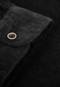 Detailaufnahme von schwarzem Cordstoff mit umgeschlagenem Kragen, der einen Metallknopf mit schwarzem Zentrum und strukturierten Oberfläche zeigt.