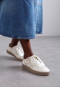 Baskets en cuir blanc avec des accents beige clair, motifs texturés, bout rond et semelles en caoutchouc plates, assorties à une jupe midi en denim.