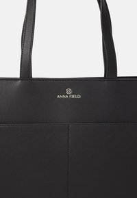 Svart tote-väska i slätt material, med två handtag, en subtil präglad logotyp och rena linjer utan synliga mönster.