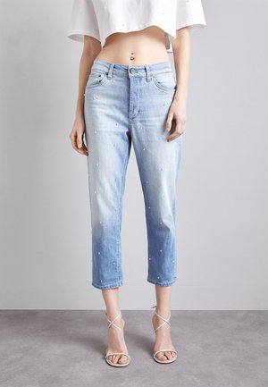 Vrouw draagt lichtblauwe cropped jeans met kleine witte versieringen, een witte cropped top en witte sandalen met bandjes, staand op een grijze vloer.