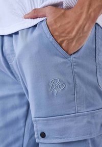 La mano di un uomo poggiata nella tasca anteriore di pantaloni cargo azzurri chiari con logo ricamato e l'orlo di una camicia bianca strutturata visibile.