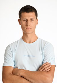 Helles blaues T-Shirt mit kurzen Ärmeln aus weichem Stoff, mit einem runden Halsausschnitt und dezent geprägtem Text "LINDBERGH" auf der Vorderseite.