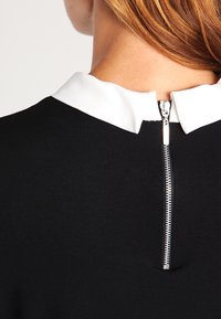 Svart plagg med en kontrasterande vit krage, försedd med en synlig bakdragkedja i metall. Slät yta.