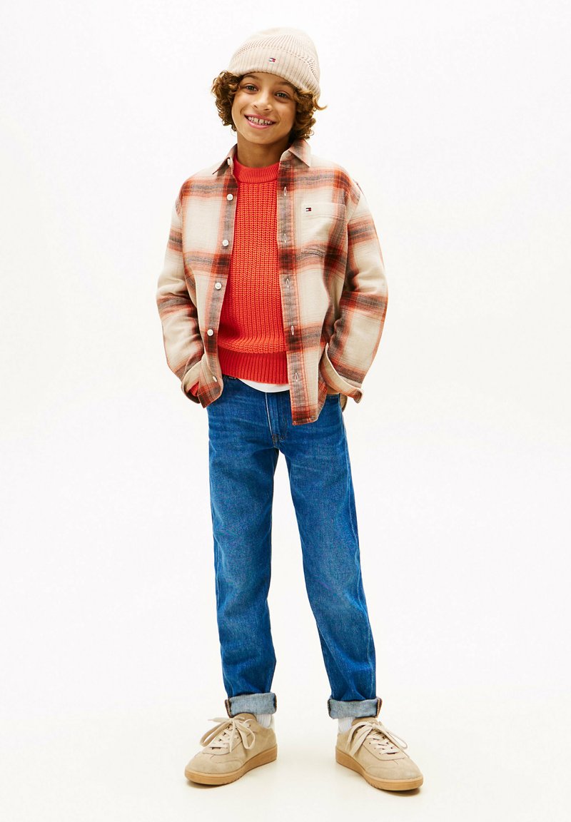 Camicia a quadri in flanella beige e arancione su un maglione rosso a maglia, abbinata a jeans blu e sneakers beige. Un berretto a maglia color crema completa il look.