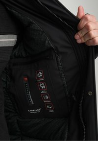 Juodas vandeniui atsparus švarkas su tekstūruota vidine dalimi, pažymėtas vėjo nepralaidumo, vandens atsparumo ir kvėpavimo savybėmis, turi užtrauktuku užsegamą kišenę viduje.