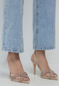 Piedi che indossano sandali trasparenti con tacchi alti e cinturini incrociati luccicanti, abbinati a jeans in denim blu chiaro tagliati sopra la caviglia.