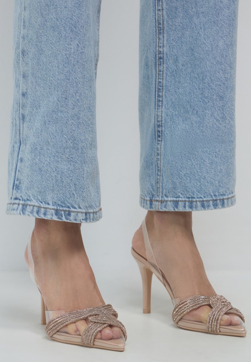 Piedi che indossano sandali trasparenti con tacchi alti e cinturini incrociati luccicanti, abbinati a jeans in denim blu chiaro tagliati sopra la caviglia.