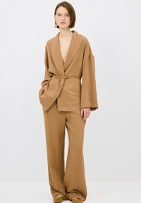 Hnědý texturovaný outfit skládající se z kabátu s hlubokým v-výstřihem a širokými rukávy, doplněný volnými kalhotami, což vytváří jednotný vzhled.
