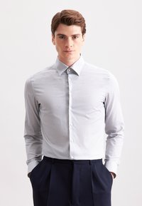 Giovane uomo che indossa una camicia a bottoni grigio chiaro con un motivo sottile e pantaloni a pieghe blu scuro, in piedi con le mani in tasca.