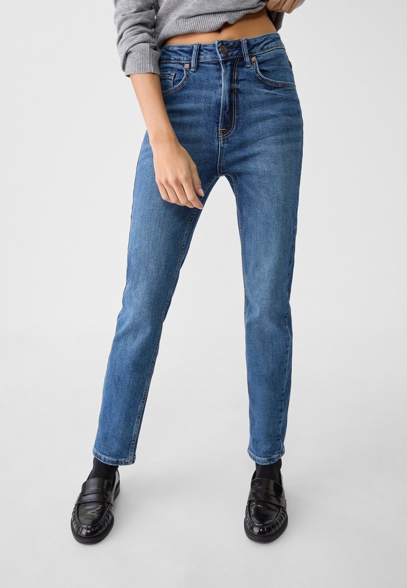 Personne portant un jean bleu taille haute, un pull gris court, des chaussettes noires et des mocassins noirs sur un fond uni.