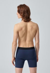 Boxers en bleu marine avec une taille élastique noire, affichant une texture lisse. Le modèle adopte une pose neutre de dos, avec les cheveux courts.