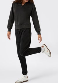 Person, der einen schwarzen Sweatshirt und Jogginghose trägt, mit weißen und braunen Sneakers, steht auf einem Bein vor einem weißen Hintergrund.