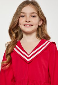 Czerwona sukienka z głębokim dekoltem w kształcie litery V i białym prążkowym kołnierzykiem. Posiada długie rękawy oraz marszczoną spódnicę. Gładki materiał o błyszczącym wykończeniu.